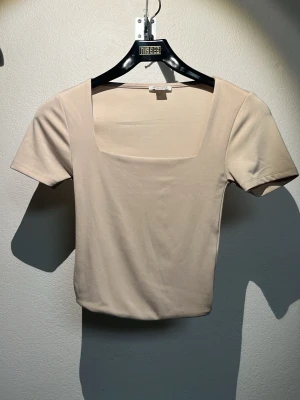 Beige topp med fyrkantig ringning Amisu - Stilren beige topp från Amisu med fyrkantig ringning och korta ärmar. Tillverkad i mjukt material som sitter snyggt och bekvämt. Perfekt att matcha med både jeans och kjol för en clean look.