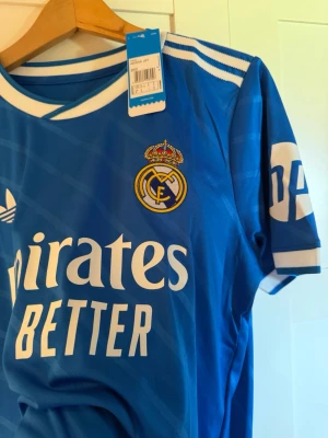 Blå Real Madrid matchtröja Adidas - Snygg blå Real Madrid matchtröja från Adidas med vita detaljer och klubbmärke på bröstet. Tröjan har klassiska Adidas-ränder på axlarna, rund halsringning och tryckt sponsorlogga framtill. Perfekt för fotbollsfans och träning.