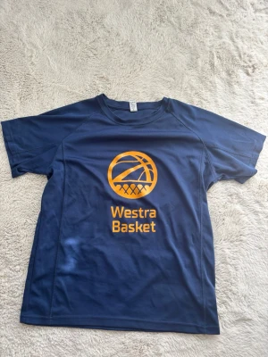 Blå Westra Basket t-shirt - Snygg blå funktions-t-shirt med gult Westra Basket-tryck framtill och 'Distriktslag' på ryggen. Perfekt för basketträning eller match. Tillverkad i ett lätt och snabbtorkande material som andas bra.