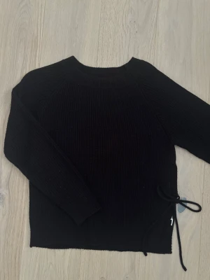 Svart stickad tröja med öppning - Snygg svart stickad tröja med rund hals och långa ärmar. Tröjan har ett unikt knytband i sidan som ger en cool detalj. Ribbstickad struktur och enkel, stilren design som passar till det mesta. Perfekt för dig som gillar minimalistisk stil.