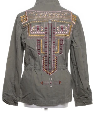 Olivgrön vindjacka med brodyr - Snygg olivgrön vindjacka med färgglad brodyr på ryggen i gult, lila och rött. Jackan har hög krage, tryckknappar framtill, två bröstfickor och dragsko i midjan för en schysst passform. Materialet är bomull och jackan har en avslappnad, trendig look.