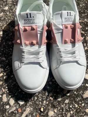 Valentino Rockstud sneakers vit/rosa - Snygga vita sneakers från Valentino med breda rosa band över vristen dekorerade med ikoniska nitar. Skorna har en klassisk låg siluett, vita snören och är tillverkade i slätt läder. Perfekta för dig som vill sticka ut med en lyxig touch.