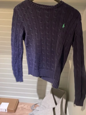 Mörklila kabelstickad tröja Polo Ralph Lauren - Kabelstickad tröja i mörklila från Polo Ralph Lauren med rund halsringning och grönt broderat logotyp på bröstet. Tröjan har långa ärmar och ribbade muddar vid ärmslut och nederkant. Perfekt för lager på lager under kalla dagar.