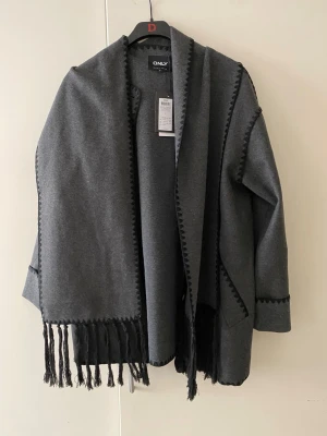 Mörkgrå kappa från ONLY med fransar - Stilren mörkgrå kappa från ONLY med dekorativa svarta sömmar och fransar längs kanten. Kappan har knappar framtill, två fickor och en lös passform. Perfekt för lager på lager och ger en cool vibe till höst- och vinterlooken.