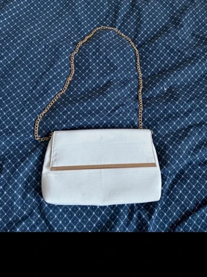 Vit kuvertväska med guldkedja - Stilren vit kuvertväska med elegant guldfärgad kedja och diskret gulddetalj på locket. Väskan har en slät, lätt glansig yta och klassisk rektangulär form. Perfekt för dig som vill ha en enkel men snygg accessoar till din outfit. Den har en liten fläck syns på sista bilden säljer den även på vinted om man föredrar att köpa där ifrån 