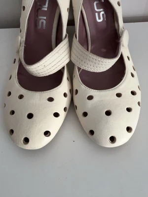 Vita pumps med hålmönster från Mjus nya - Unika vita pumps från Mjus  med rund tå och dekorativa hål över hela skon. Skorna har en bred rem med knappstängning över foten och en mjuk, matt finish. Perfekta för dig som vill sticka ut med en retroinspirerad look.