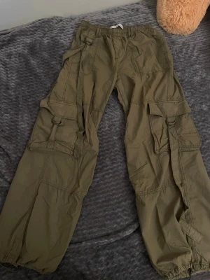 Bershka Olivgröna cargobyxor med fickor - Säljer ett par olivgröna cargobyxor med flera stora fickor och coola detaljer som D-ringar och justerbara benslut. Byxorna har avslappnad passform och är perfekta för en streetwear-look. Materialet känns lätt och bekvämt, perfekt för dig som gillar utility-stil.