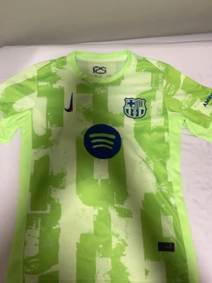 Barcelona matchtröja limegrön Nike - Säljer en limegrön Barcelona matchtröja från Nike med blå detaljer, klubbmärke och Spotify-logga på bröstet. Nummer 10 och Lamine Yamal på ryggen samt UNHCR-logga. Tröjan har korta ärmar och modern design, perfekt för fotbollsträning eller support.