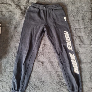 Svarta mjukisbyxor Billie Eilish - Svarta mjukisbyxor från Billie Eilish med tryckt logga i vitt längs ena benet. Byxorna har elastisk midja och muddar vid bensluten för skön passform. Perfekta för chill eller träning, tillverkade i mjukt bomullsmaterial.