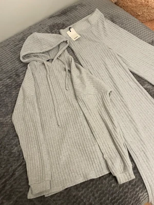 Grått ribbat loungewear-set med huva - Mysigt loungewear-set i ljusgrått med ribbad struktur. Setet består av en hoodie med dragsko och långa ärmar samt matchande vida byxor med resår i midjan. Perfekt för chill dagar hemma eller när du vill vara extra bekväm.
