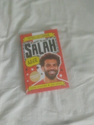 Fotbollsstjärnor: Salah äger - En färgstark bok om fotbollsstjärnan Salah, skriven av Simon Mugford och Dan Green. Omslaget är rött med svartvit text och illustrationer. Inuti finns svartvita illustrationer och lättläst text, perfekt för unga som gillar sport och inspirerande berättelser.