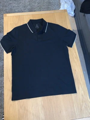 L. Mango pikétröja med vit kant - Bara en vecka gammal köpte till min lille bror va lite stor han har M var försent och returnera efter borttagen prislapp Snygg svart pikétröja från mango med klassisk krage och vita detaljer längs krage och ärmslut. Tröjan är kortärmad perfekt för en stilren och enkel look. Är svart men ser utt som marinblå av belysning