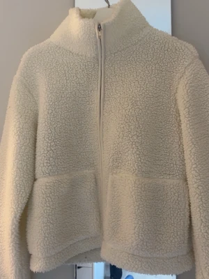 Vit teddyjacka med dragkedja - Mysig vit teddyjacka i fluffigt material med hög krage och hel dragkedja framtill. Jackan har två stora fickor fram och en avslappnad passform. Perfekt för att hålla värmen under kyliga dagar. Materialet är mjukt och ger en trendig look.
