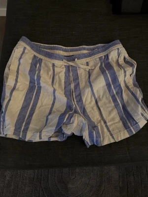 Randiga blåvita shorts med snörning - Säljer ett par avslappnade shorts med blå och vita ränder. Shortsen har elastisk midja med snörning och en bakficka. Perfekta för varma dagar och har en luftig passform. Materialet känns mjukt och svalt, linnen. 