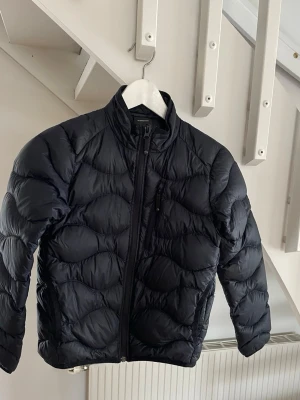 Svart quiltad pufferjacka - Snygg svart pufferjacka med quiltat mönster och hög krage. Jackan har dragkedja framtill och en bröstficka med dragkedja. Perfekt för kyliga dagar och ger en modern look. Den har ett litet hål på vänstra armen men den är lagad och syns int