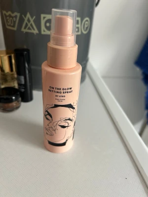 On The Glow Setting Spray från Lyko - On The Glow Setting Spray från Lyko är en face mist i en ljusrosa plastflaska med svart minimalistisk ansiktsillustration. Sprayen ger en dewy finish och kommer i en smidig pumpflaska på 100 ml, perfekt för att fräscha upp och fixera makeupen under dagen.