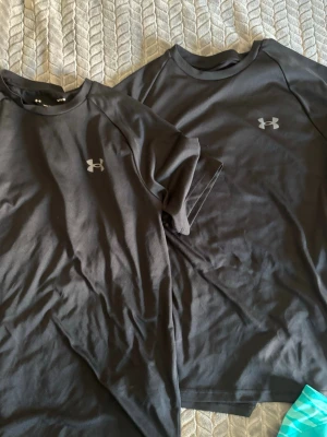 Svarta Under Armour t-shirts - Två svarta t-shirts från Under Armour med klassisk logga på bröstet och en T-shirt från Nike, alla är dry fit och man får alla 3 tillsammans. T-shirtsen har rund hals, korta ärmar och är tillverkade i ett mjukt, funktionellt material som passar perfekt till träning. Snygg och enkel design som funkar till det mesta.