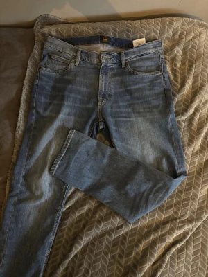 Blå slim fit jeans från Lee - Säljer ett par klassiska blå slim fit jeans från Lee. Jeansen har fem fickor, normal midja och dragkedjegylf. Materialet är mjukt denim i bomull med en snygg tvättad look och subtila slitningar. Perfekta till vardagsstilen! Storlek 33W/30L