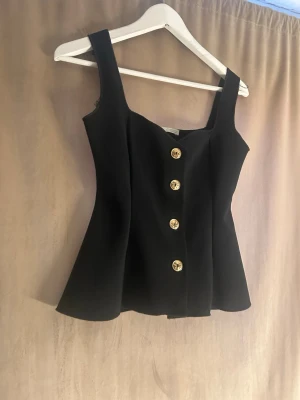 Svart korsettopp med guldknappar - Stilren svart korsettopp med breda axelband och dekorativa guldknappar framtill. Toppen har en figurnära passform och lätt utsvängd nederkant som ger en snygg siluett. Perfekt att styla med jeans eller kjol för en trendig look.