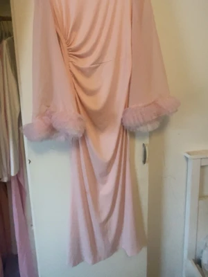 Ljusrosa långklänning med volangärm - Säljer en ljusrosa långklänning med vida, långa ärmar och volang i tyll vid ärmsluten. Klänningen har draperad front och ett mjukt, flytande tyg som ger en romantisk och unik look. Perfekt för dig som gillar feminina detaljer och statement-ärmar.