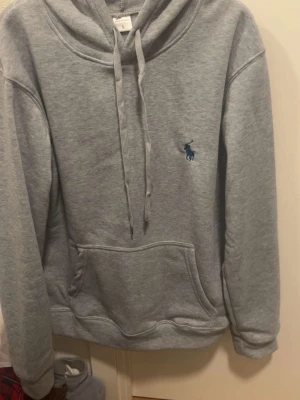 Grå hoodie från Polo Ralph Lauren - Snygg grå hoodie från Polo Ralph Lauren med klassisk blå broderad logga på bröstet. Hoodien har huva med dragsko, stor magficka och mjukt material som känns riktigt skönt. Perfekt för en chill och avslappnad stil.