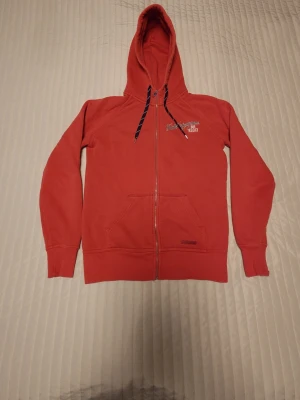 Röd hoodie från Peak Performance - Snygg röd hoodie från Peak Performance med dragkedja, huva med snören och känguruficka framtill. Tillverkad i mjukt material som känns skönt mot huden. Diskret logga på bröstet och klassisk passform. Perfekt för en avslappnad och sportig stil.