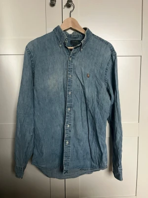Blå jeansskjorta från Polo Ralph Lauren - Klassisk blå jeansskjorta från Polo Ralph Lauren med broderad logga på bröstet. Skjortan har lång ärm, knappar framtill och en avslappnad passform. Perfekt till jeans eller chinos för en casual look.