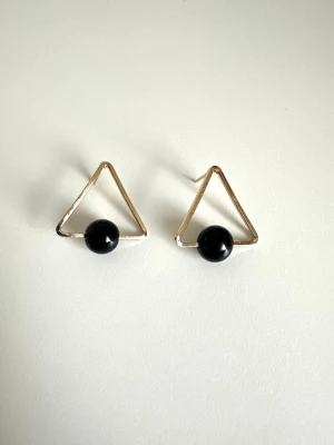 Guldiga triangelformade örhängen, helt NYA - Helt NYA snygga örhängen med guldiga trianglar och svarta pärlor. De har en modern och minimalistisk design som verkligen sticker ut. Perfekta för dig som gillar geometriska former och stilrena accessoarer. Materialet är metall och plastpärla. Säljes helt NYA i förpackning. 
