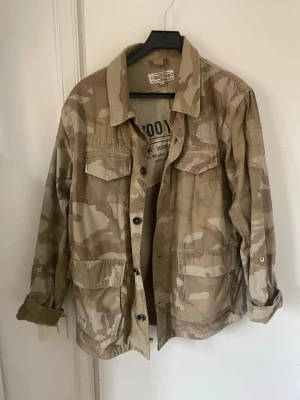 Klassisk camo overshirt - Säljer en overshirt i klassiskt beige och brunt camomönster. Skjortan har fyra stora fickor framtill, knäppning med svarta knappar och en avslappnad passform. Tillverkad i slitstark bomull, perfekt för lager på lager och streetwear-stil.