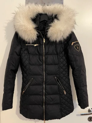 Svart dunjacka Rockandblue med pälshuva - Snygg svart dunjacka från Rockandblue med stor fluffig pälskant på huvan och guldfärgade dragkedjor. Jackan har quiltade detaljer, två snedställda fickor med dragkedja och en bröstficka. Perfekt för kalla vinterdagar och riktigt trendig look.