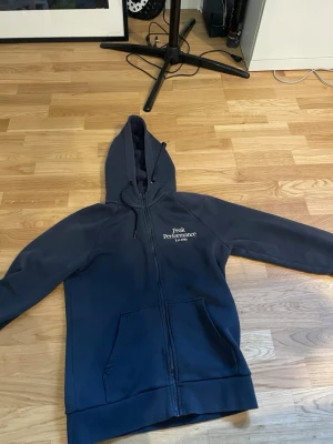 Mörkblå hoodie från Peak Performance - Snygg mörkblå hoodie från Peak Performance med dragkedja framtill och två fickor. Märkets logga är broderad på bröstet. Hoodien har en klassisk huva med dragsko och ribbade muddar vid ärmslut och nederkant. Perfekt för en avslappnad stil.