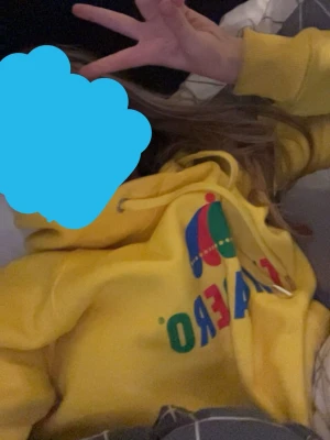 Gul Trocadero hoodie med tryck - Säljer en gul hoodie från Trocadero med färgglatt tryck på bröstet och klassisk huva med dragsko. Hoodien har känguruficka framtill och ribbade muddar vid ärmslut och nederkant. Perfekt för dig som gillar streetwear och vill sticka ut med en ikonisk svensk design.