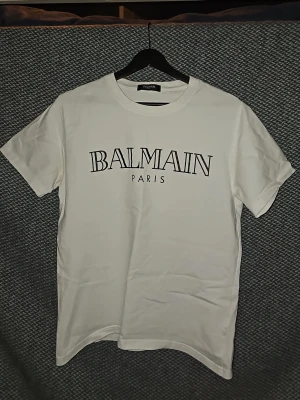 Vit Balmain Paris t-shirt - Säljer en clean vit t-shirt från Balmain Paris med stor svart logga framtill. Klassisk rund halsringning och korta ärmar. Perfekt basic-plagg för dig som gillar exklusiva märken och enkel stil. Är öppen för prisförslag vid snabb försäljning.