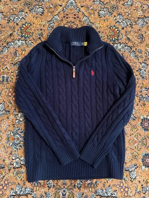 Mörkblå kabelstickad tröja Polo Ralph Lauren - Mörkblå kabelstickad tröja från Polo Ralph Lauren med halv dragkedja och hög krage. Tröjan har ett rött broderat logomärke på bröstet och ribbade muddar. Perfekt för dig som gillar klassisk och stilren look. Den passa jätte bra för både M och S