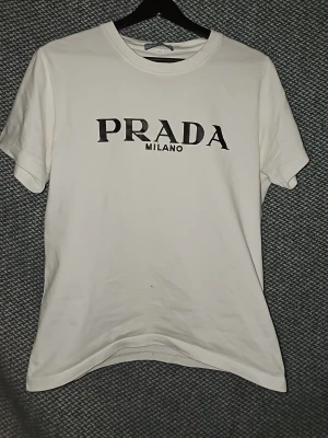 Vit t-shirt från Prada Milano - Snygg vit t-shirt från Prada Milano med klassisk svart logga framtill. T-shirten har rund halsringning och korta ärmar. Tillverkad i mjuk bomull för en skön och avslappnad look. Är öppen för prisförslag vid snabb försäljning.