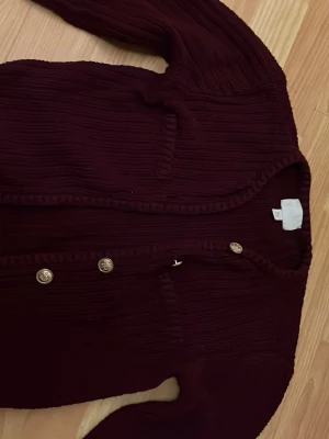 Vinröd ribbad kofta med guldknappar - Mysig vinröd kofta med ribbad struktur och dekorativa guldknappar framtill. Koftan har v-ringning, långa ärmar och två fickor på framsidan. Perfekt för lager på lager och en snygg detalj till din outfit.