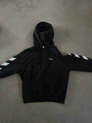Svart hoodie från Off-White - Säljer en svart hoodie från Off-White med vita diagonala ränder på ärmarna och liten logga på bröstet. Klassisk känguruficka framtill och justerbar huva med snören. Perfekt för dig som gillar streetwear och vill sticka ut lite extra. Liten S men lång i armarna 