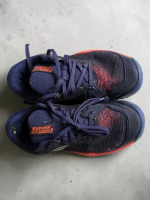 Babolat Jet Mach 3 mörkblå sneakers - Säljer ett par Babolat Jet Mach 3 sneakers i mörkblå mesh med röda detaljer och orange logga på plösen. Skorna har snörning, slitstark sula och sportig design, perfekta för tennis eller padel. Lätta och ventilerande material för maximal komfort på banan.