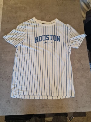 Vit randig Houston t-shirt - Snygg vit t-shirt med blå vertikala ränder och texten 'HOUSTON AMERICA' på bröstet. Klassisk passform och rund halsringning. Perfekt för dig som gillar sportig och avslappnad stil.