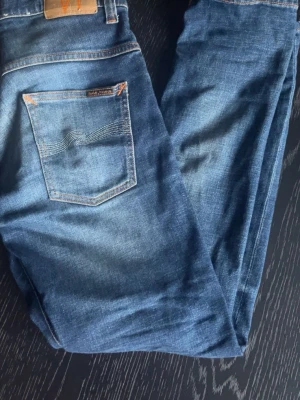 Blå jeans från Nudie Jeans, 30/30 - Säljer ett par klassiska blå jeans från Nudie Jeans i storlek 30/30. Jeansen har en snygg tvätt, raka ben och kontrastsömmar. Tillverkade i slitstarkt denim med en bakficka som har Nudie Jeans signaturbrodyr.
