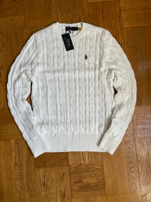 Vit kabelstickad tröja Polo Ralph Lauren - Kabelstickad vit tröja från Polo Ralph Lauren med klassisk rund halsringning och marinblå broderad logga på bröstet. Tröjan har ribbade muddar vid ärmslut och nederkant. Perfekt för dig som gillar stilrena och tidlösa plagg.