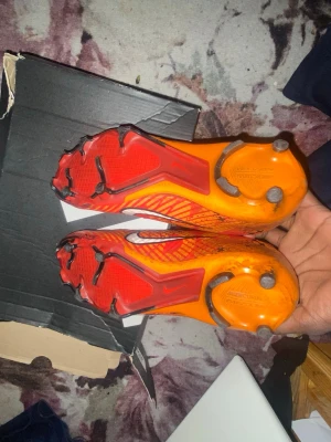 Nike orangea Mercurial fotbollsskor - Säljer ett par Nike Mercurial fotbollsskor i en riktigt snygg orange färg med röda och svarta detaljer. Skorna har en vit Nike-logga på sidan och är designade med fasta dobbar för bra grepp på planen. Ovandelen är i syntetmaterial för lätt vikt och bra bollkontroll.