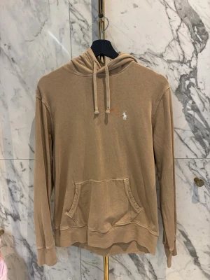 Beige hoodie från Polo Ralph Lauren - Snygg beige hoodie från Polo Ralph Lauren med klassisk känguruficka och dragsko i huvan. Liten vit broderad logga på bröstet. Tillverkad i mjukt bomullsmaterial och har ribbade muddar vid ärmslut och nederkant. Perfekt för en chill och avslappnad stil.