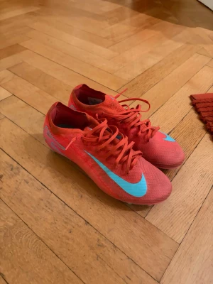 Nike Mercurial röda fotbollsskor - Säljer ett par Nike Mercurial fotbollsskor i rött med ljusblå swoosh och detaljer. Skorna har snörning och är tillverkade i ett lätt syntetmaterial för bra bollkontroll och grepp på planen. Perfekta för dig som vill ha speed och stil på matchen. Lite smutsiga och limmet har släppt lite på höger tå, men inget som man tänker på när man spelar! 