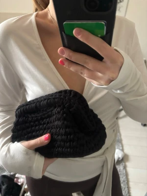 Svart virkad kuvertväska - Snygg svart kuvertväska i virkat material. Väskan har en mjuk, rektangulär form och stängs upptill. Perfekt att bära i handen och ger en trendig touch till din outfit. Märke saknas.