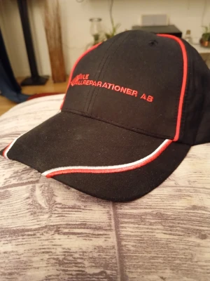 Svart keps med röd och vit detalj - Snygg svart keps med böjd skärm och broderad text i rött framtill. Kepsen har röda och vita detaljer längs skärmen och panelerna. Justerbar baktill för perfekt passform. Perfekt accessoar för en sportig och avslappnad stil.