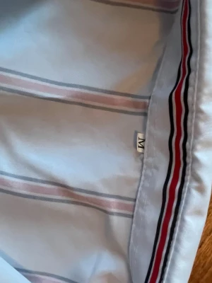 Vit randig skjorta med röda detaljer - Säljer en vit skjorta med tunna röda och svarta ränder. Skjortan har klassisk krage, knäppning framtill och är tillverkad i ett mjukt bomullstyg. Perfekt för dig som gillar stilrena och fräscha plagg med lite färg.