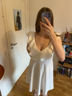 Studentklänning - Supersöt vit klänning med volangdetaljer längs axlar och urringning. Klänningen har en v-ringad front och är ärmlös med en lätt utsvängd kjol. Lappen är bortklippt i ryggen men den är från bubbleroom i storlek M 