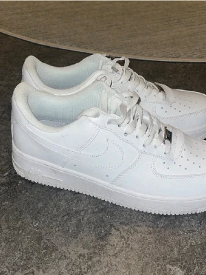 Vita Nike Air Force 1 sneakers - Klassiska helvita Nike Air Force 1 sneakers i lågt utförande. Skorna har en stilren design med perforerad tå, vit sula och Nike swoosh på sidan. Tillverkade i läder med vita skosnören och diskret branding på plösen.