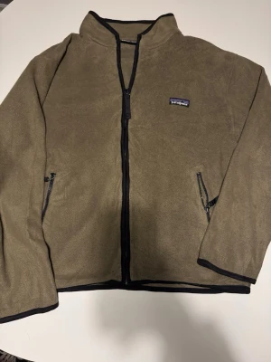 Patagonia fleecejacka khaki/olivgrön , unisex - Säljer en olivgrön fleecejacka från Patagonia med  dragkedja framtill och svart kantband. Tillverkad i mjukt fleecematerial, perfekt för lager-på-lager. Mått: längd 62cm, bred armhåla till armhåla 57cm. Patagonia-logga på bröstet ger en clean och stilren look.